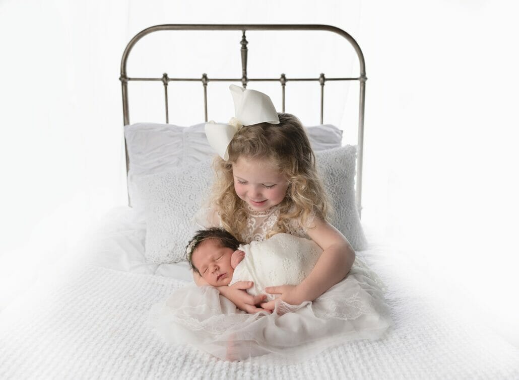 A little girl holds a newborn baby on a white bed in Baton Rouge newborn photography.