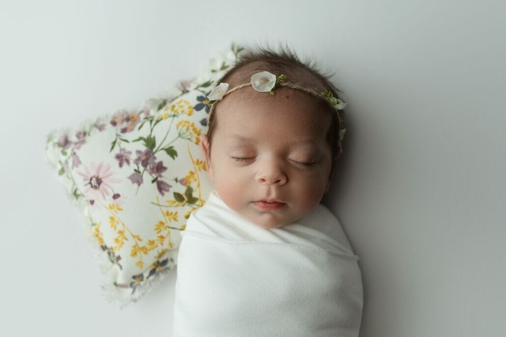 A newborn photo of a swaddled baby with her head on a floral pillow