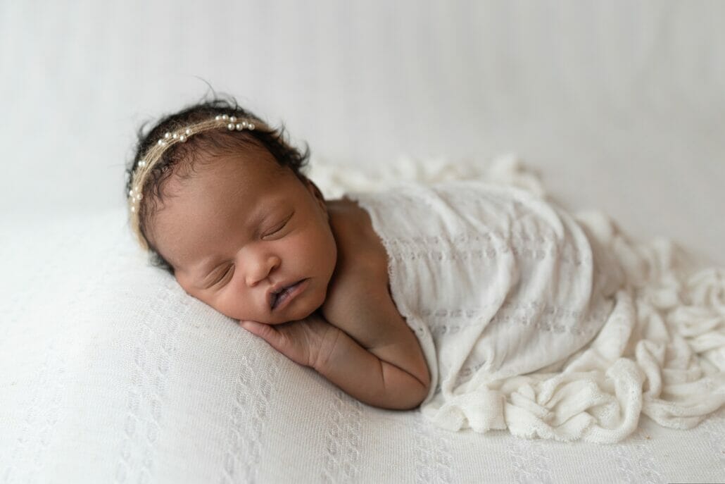 A newborn baby lays on a white backdrop with a blanket draped over her for posing for newborn photos.