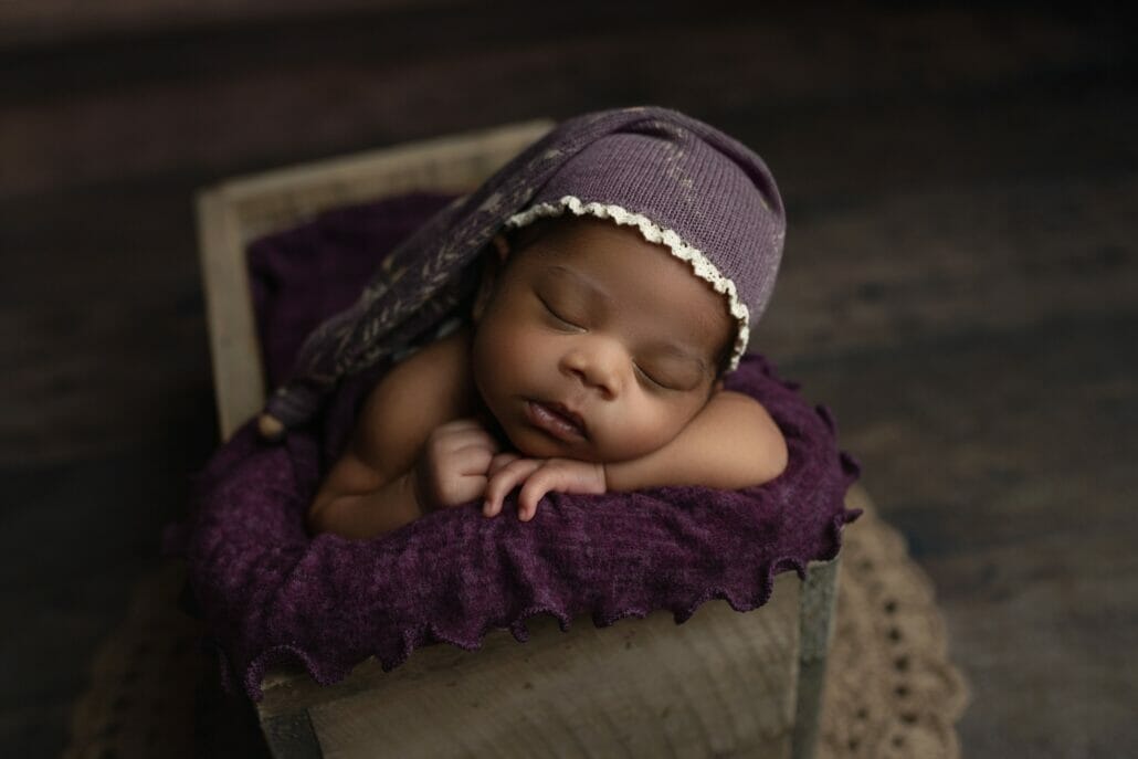A newborn baby is posed in a basket wearing a purple hat.