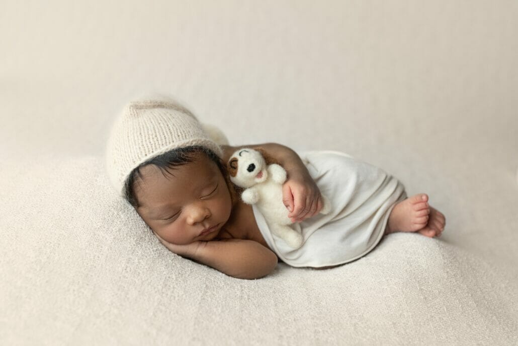 A baby is posed in a white blanket and cap holding a stuffy for Baton Rouge newborn photography.