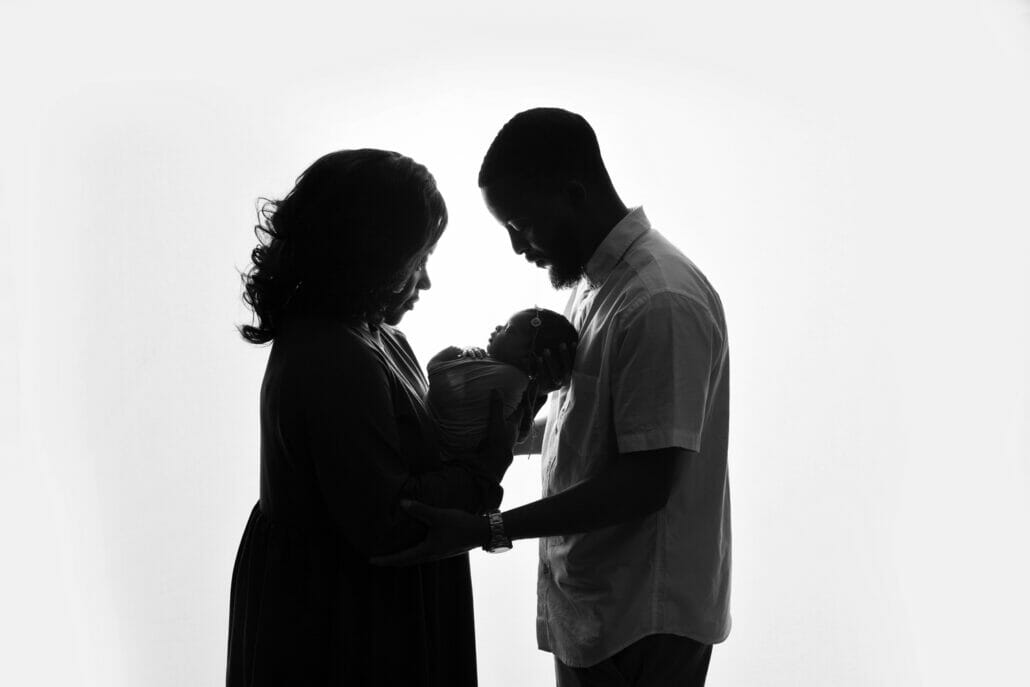 Two parents holding a newborn are silhouetted against a studio backdrop.