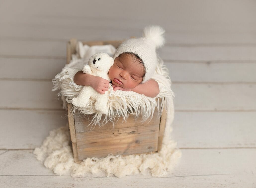 A newborn baby poses with a teddybear in a wooden crib.