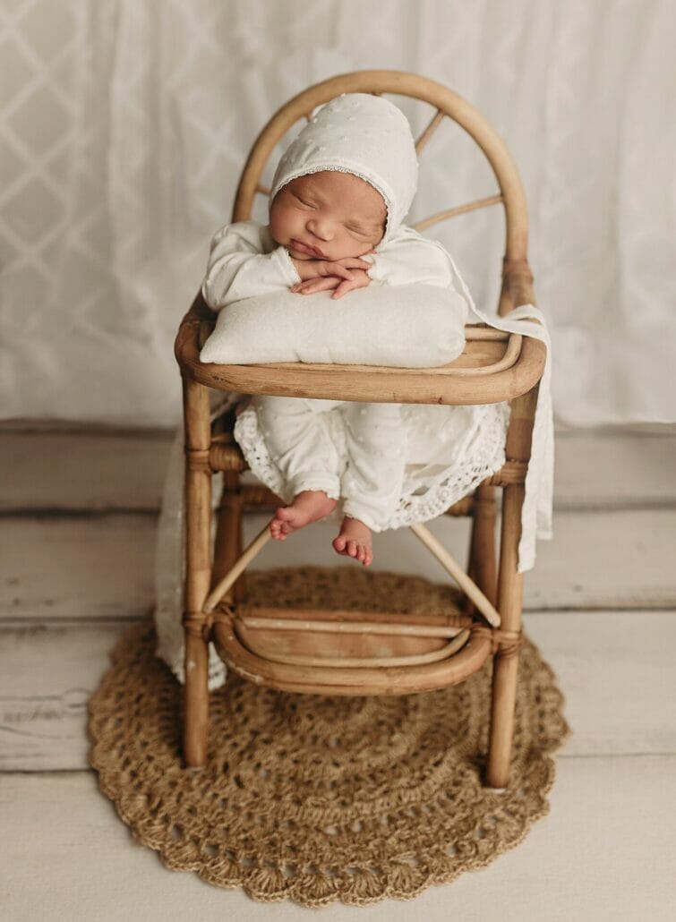 A baby sits in a wooden high chair leaning on a pillow in a newborn photography pose.