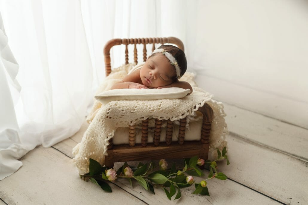 A newborn photo shows a baby on a tiny wooden bed in a photography studio.