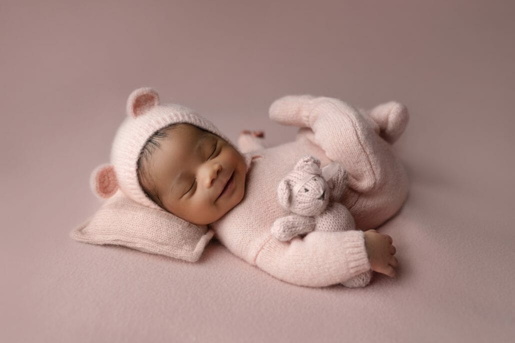A baby in a pink onesie holds a teddy bear.