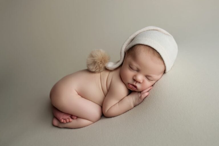 A newborn baby sleeps on their side on a soft, light-colored surface, wearing a cream-colored knit hat with a fluffy pom-pom. The baby’s hands are tucked under their head and their legs are curled up. Melinda Gilmore Photography - Maternity & Newborn Photographer - Louisiana