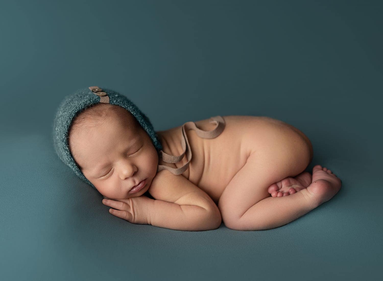 A newborn baby sleeps peacefully on a teal background, lying on their side with hands under their cheek, wearing a soft teal bonnet and a neutral-colored outfit. Melinda Gilmore Photography - Maternity & Newborn Photographer - Louisiana
