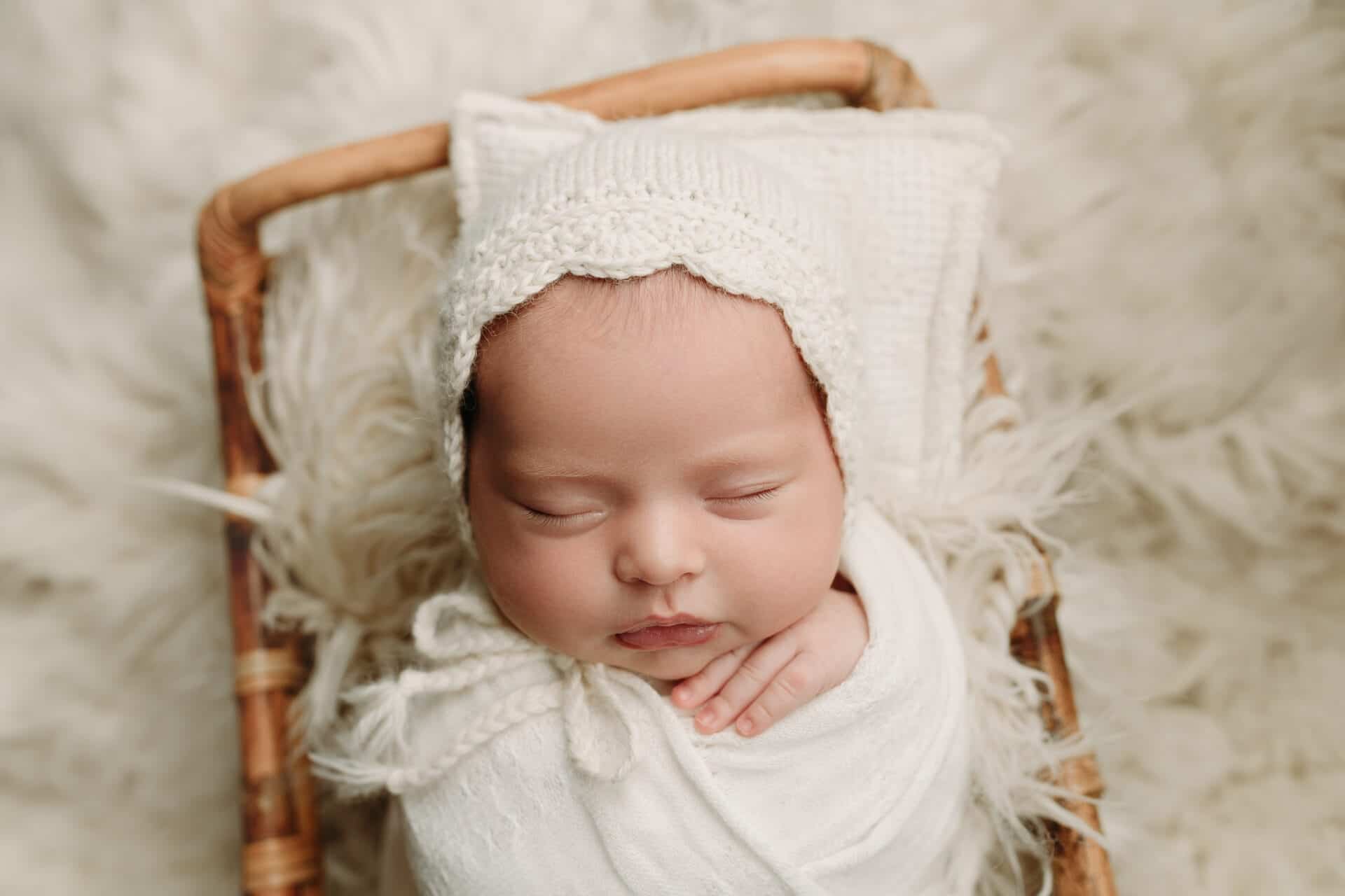 A newborn baby sleeps peacefully, wrapped in a white blanket and wearing a knitted white bonnet, lying on a soft, furry surface with a small woven pillow and bassinet beneath them. Melinda Gilmore Photography - Maternity & Newborn Photographer - Louisiana