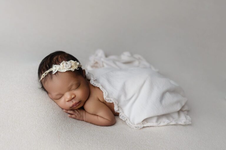 A newborn baby sleeps peacefully on a soft white blanket, with a delicate cream lace headband and a light blanket partially covering their body. The background is neutral and softly lit. Melinda Gilmore Photography - Maternity & Newborn Photographer - Louisiana