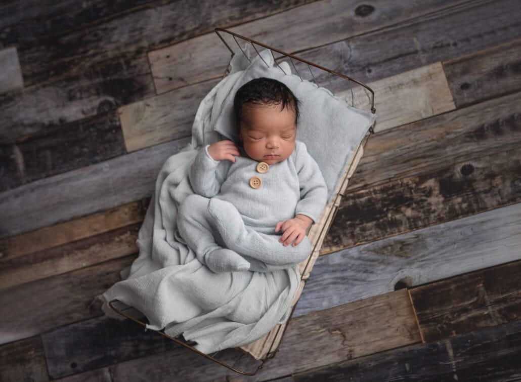 The newborn boy is in a blue onesie outfit in a tiny bed prop with a top Lafayette pediatrician.