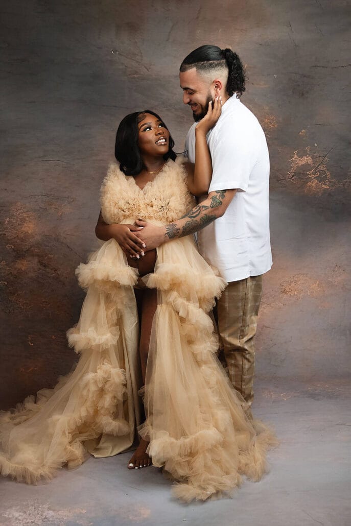 Expecting parents stand in a studio holding the bump in a cream tule dress and white polo after meeting new orleans doulas