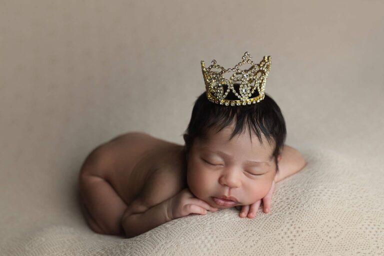 A newborn baby sleeps on a soft beige blanket, resting their head on their hands. The baby has dark hair and wears a small, decorative gold crown atop their head. Melinda Gilmore Photography - Maternity & Newborn Photographer - Louisiana
