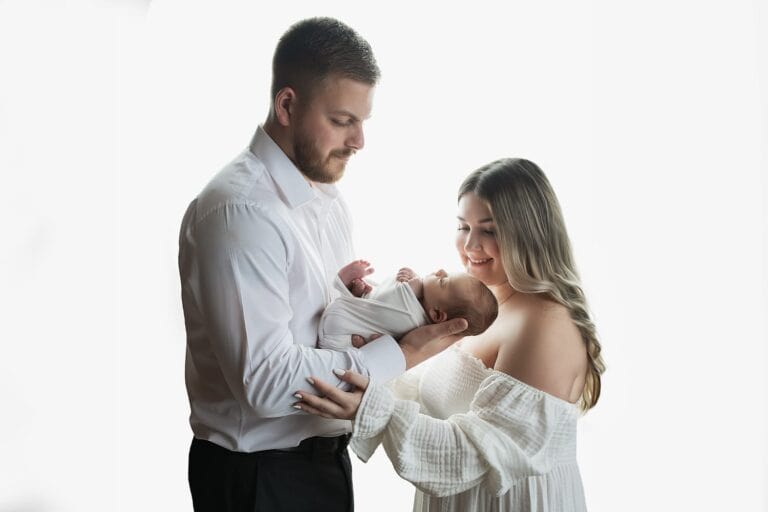A man and woman stand together, both wearing white, gently holding a newborn baby wrapped in a blanket. The baby is cradled between them, and both adults look at the baby with loving expressions against a white background. Melinda Gilmore Photography - Maternity & Newborn Photographer - Louisiana