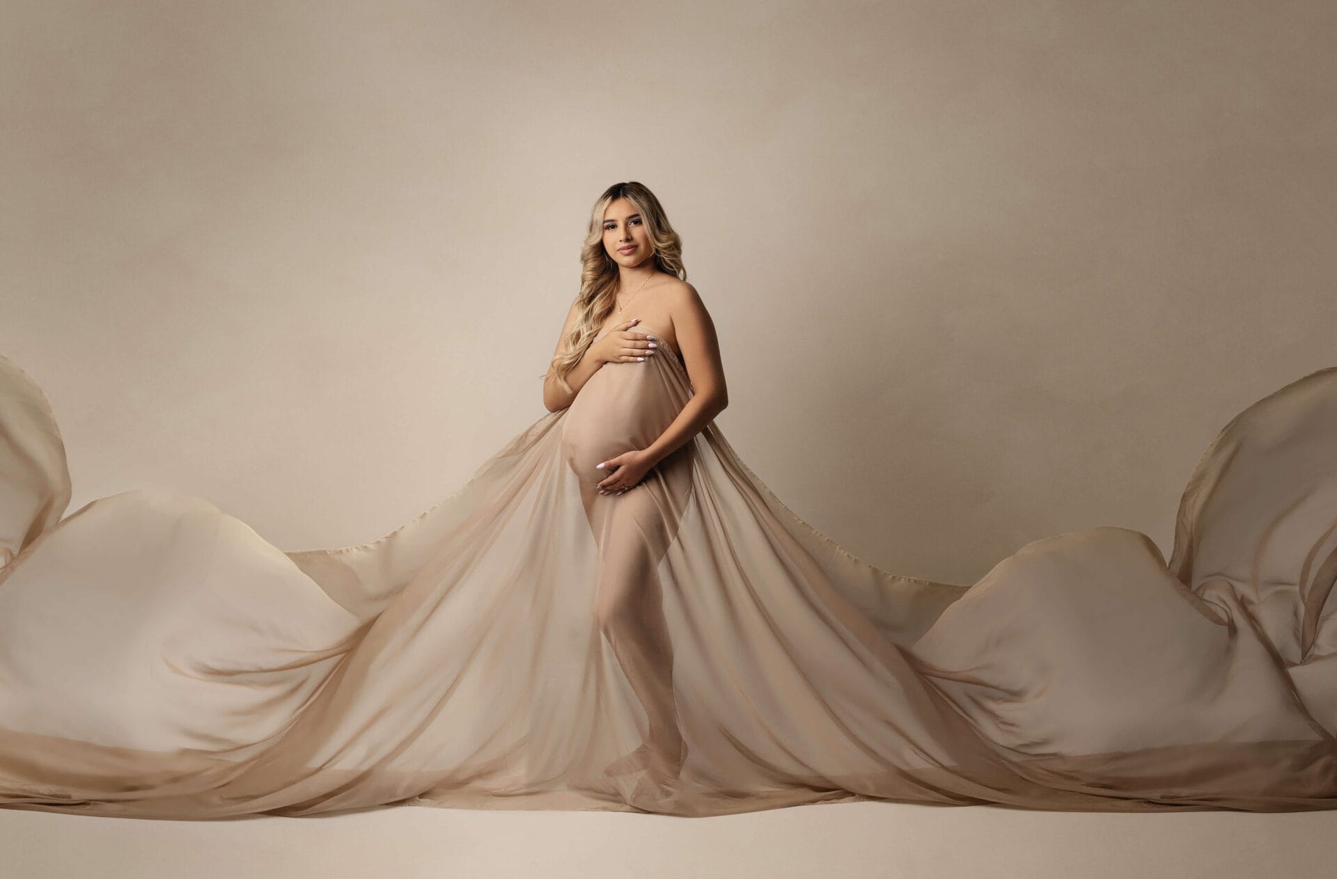A pregnant mom is to be in the studio with fabric draped, embracing her baby bump.