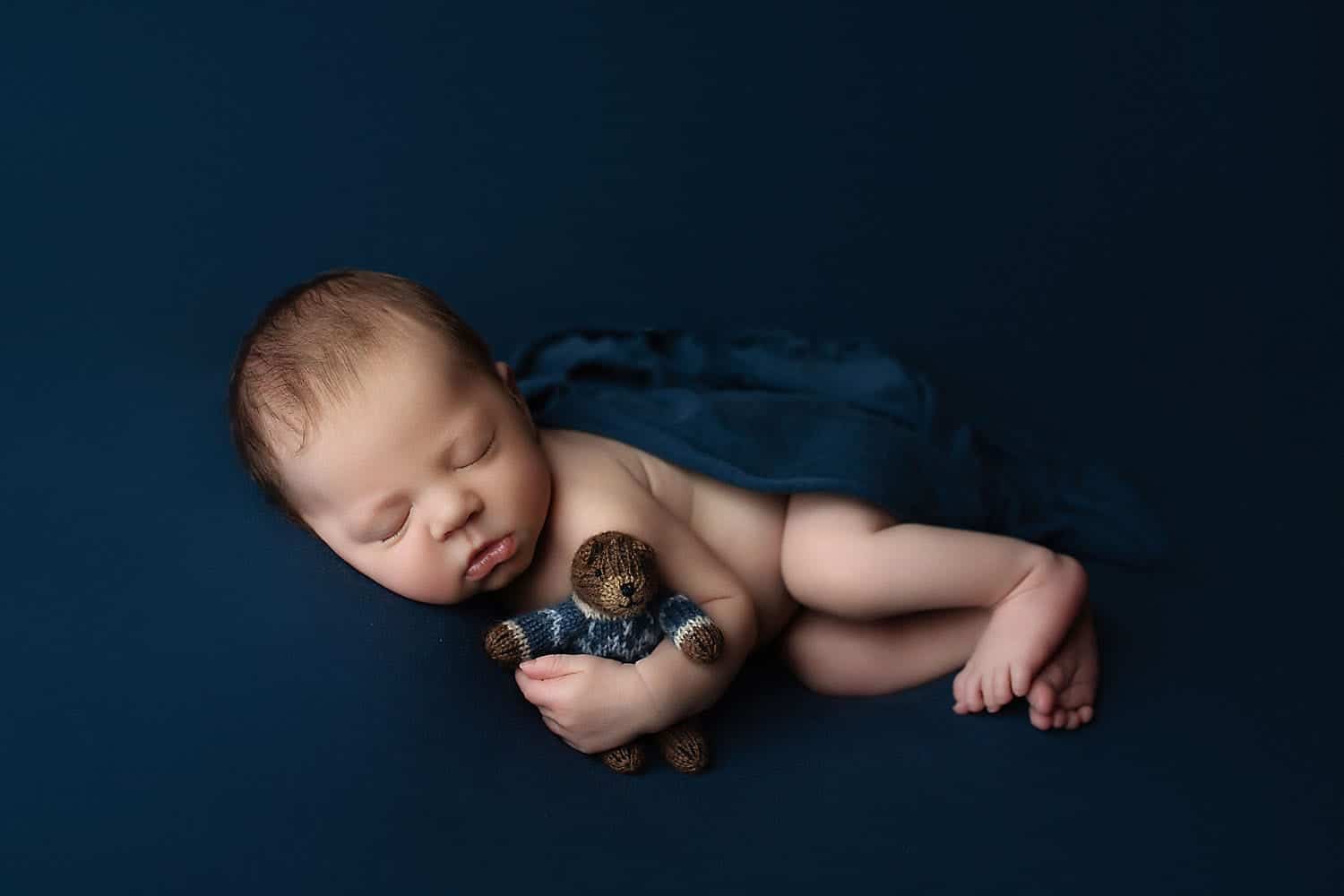 A newborn in the studio is lying on a blue backdrop, cuddling a teddy bear.
