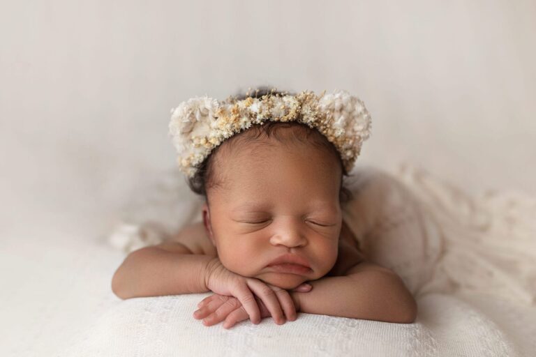 A newborn baby sleeps peacefully on a soft white blanket, resting their head on their hands. The baby wears a delicate knitted headband with bear ears, and the background is softly blurred in neutral tones. Melinda Gilmore Photography - Maternity & Newborn Photographer - Louisiana
