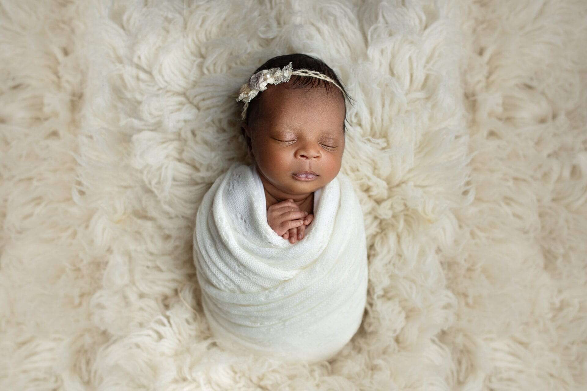 A newborn baby wrapped in a soft white blanket sleeps peacefully on a fluffy cream-colored rug, wearing a delicate floral headband. Melinda Gilmore Photography - Maternity & Newborn Photographer - Louisiana