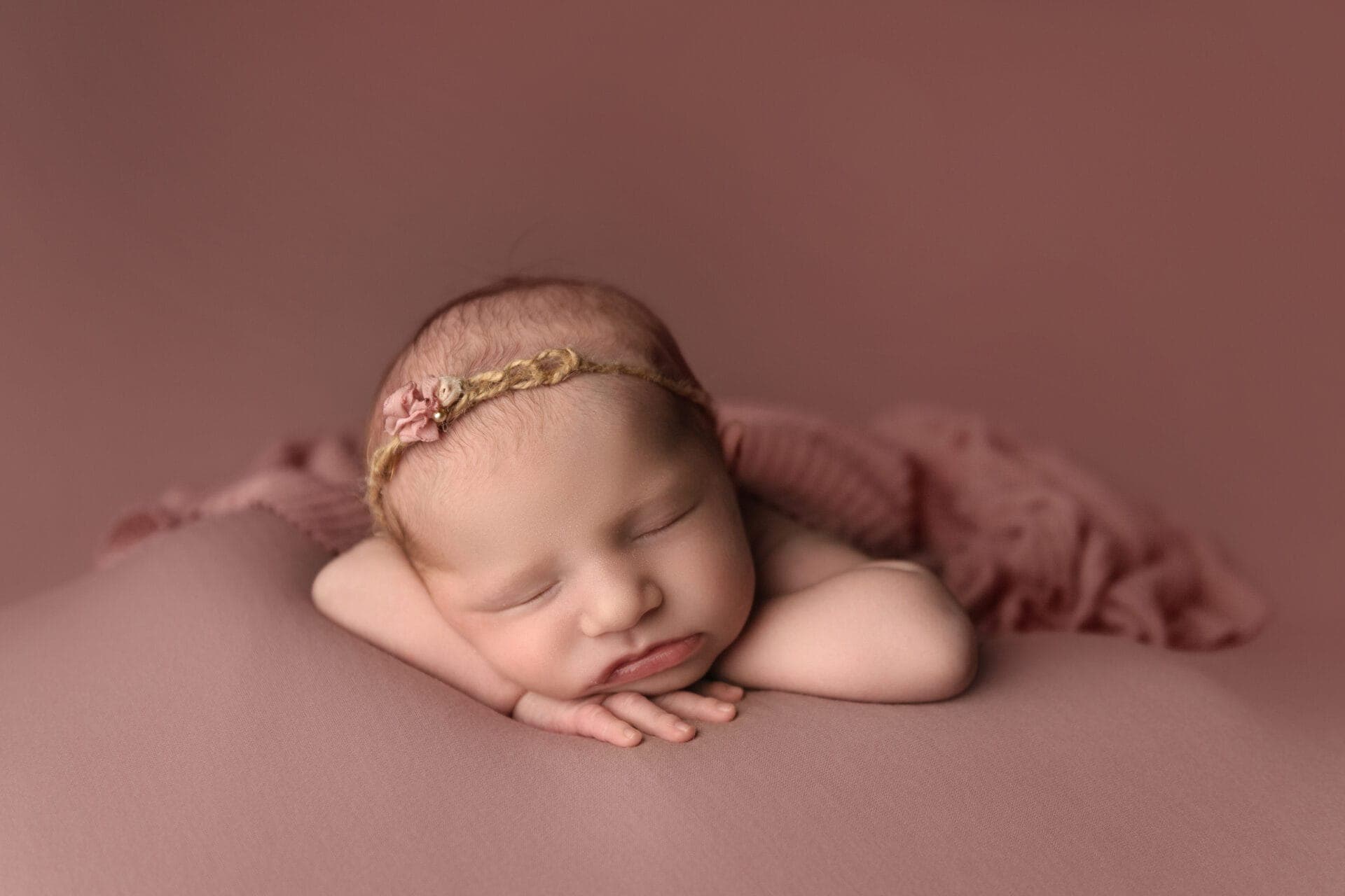 A sleeping newborn baby rests on a soft pink surface, with their head propped on their arms. The baby wears a delicate braided headband with a small flower, and is wrapped in a light pink blanket. Melinda Gilmore Photography - Maternity & <a href=