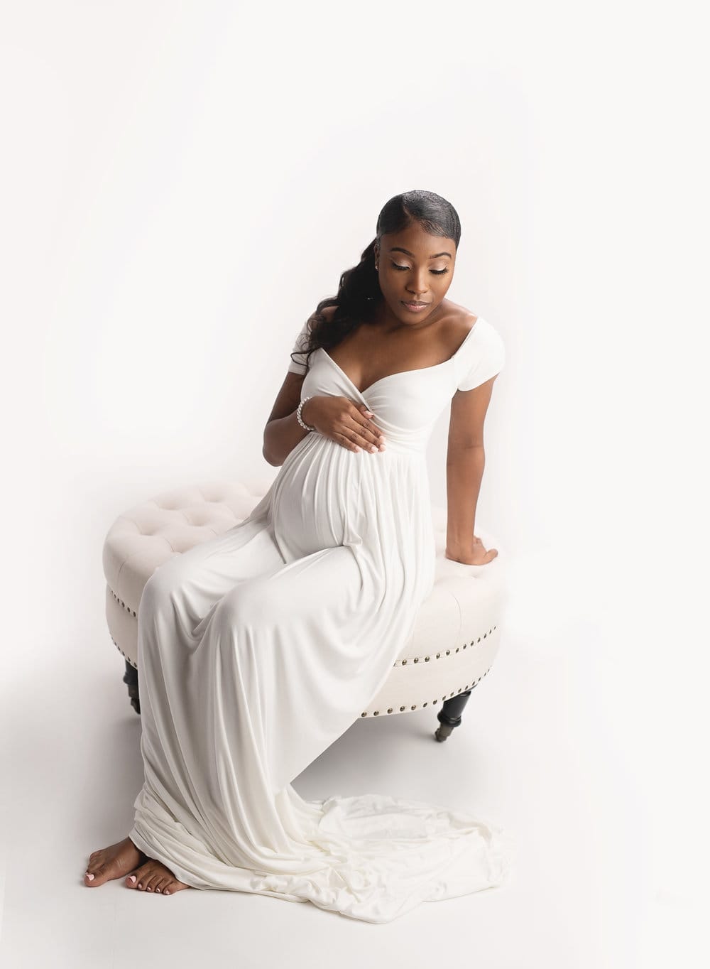A mom to be in the studio with a white gown sitting on a ottoman embracing her pregnant belly at New orleans yoga studio.