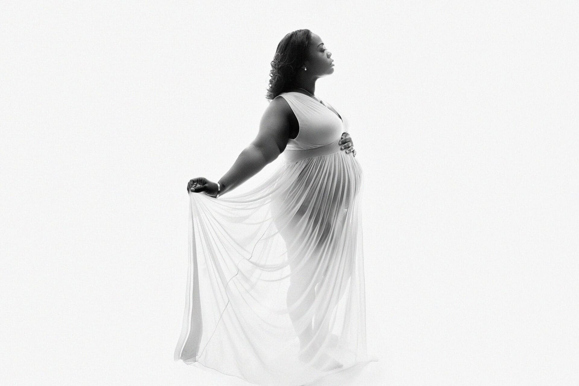 A pregnant woman in a flowing, sleeveless white dress stands in profile, gently holding the fabric of her dress. She looks upward with a serene expression against a white background. Melinda Gilmore Photography - Maternity & Newborn Photographer - Louisiana