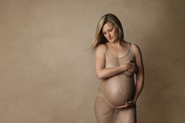 A pregnant woman with long blonde hair stands against a plain beige background, wearing a sheer dress and gently cradling her belly with one hand while covering her chest with the other, looking down thoughtfully. Melinda Gilmore Photography - Maternity & Newborn Photographer - Louisiana