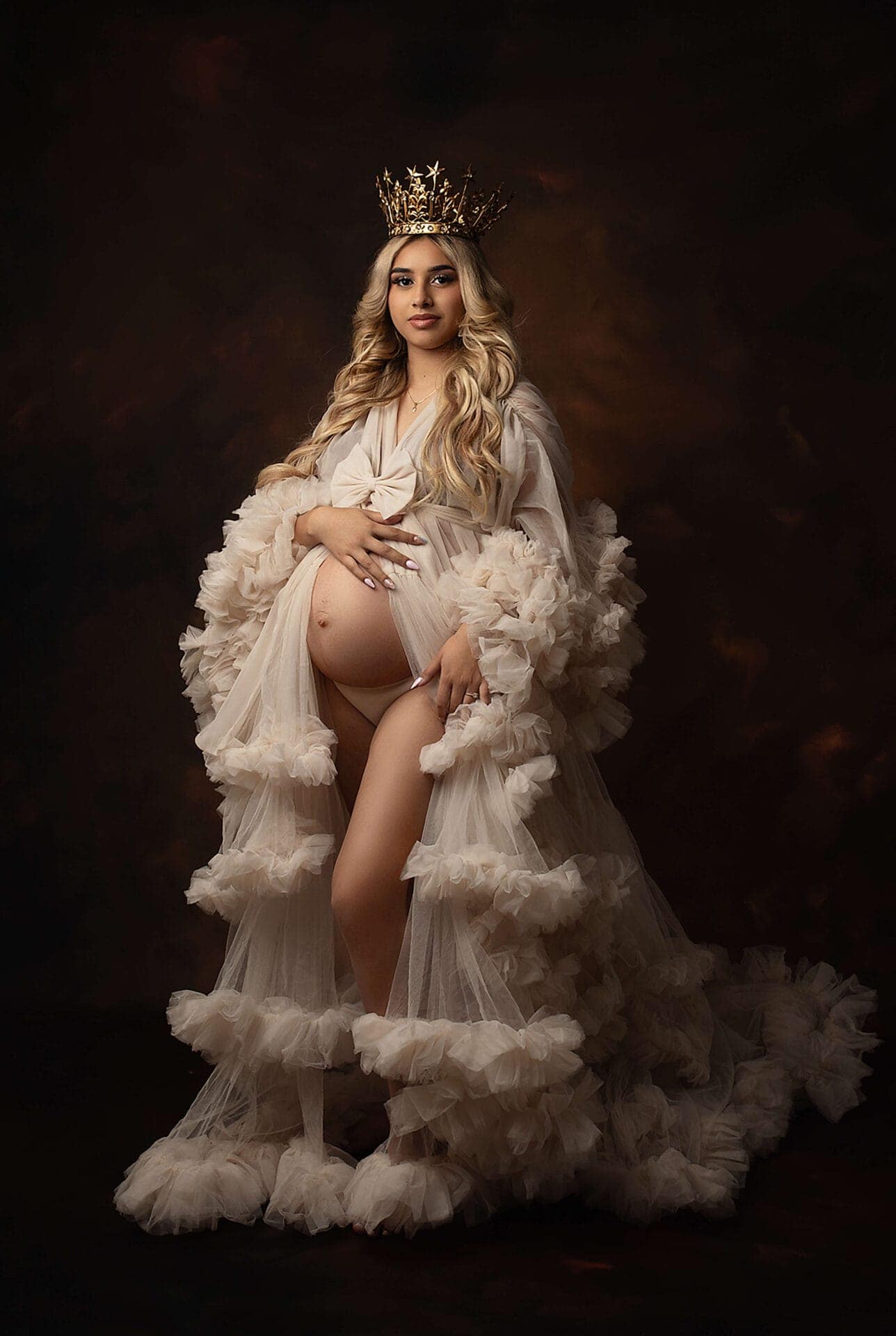 A pregnant woman with long blonde hair wears a gold crown and a sheer, ruffled, flowing robe. She stands barefoot against a dark background, cradling her belly and looking confidently at the camera. Melinda Gilmore Photography - Maternity & Newborn Photographer - Louisiana
