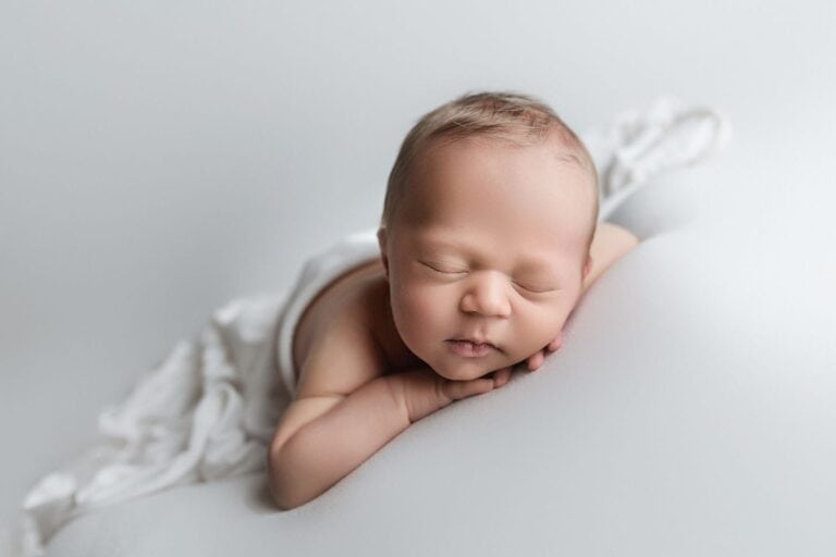 A newborn baby sleeps peacefully on a soft, white surface with their head resting on their hands, wrapped in a white blanket, against a light, neutral background. Melinda Gilmore Photography - Maternity & Newborn Photographer - Louisiana