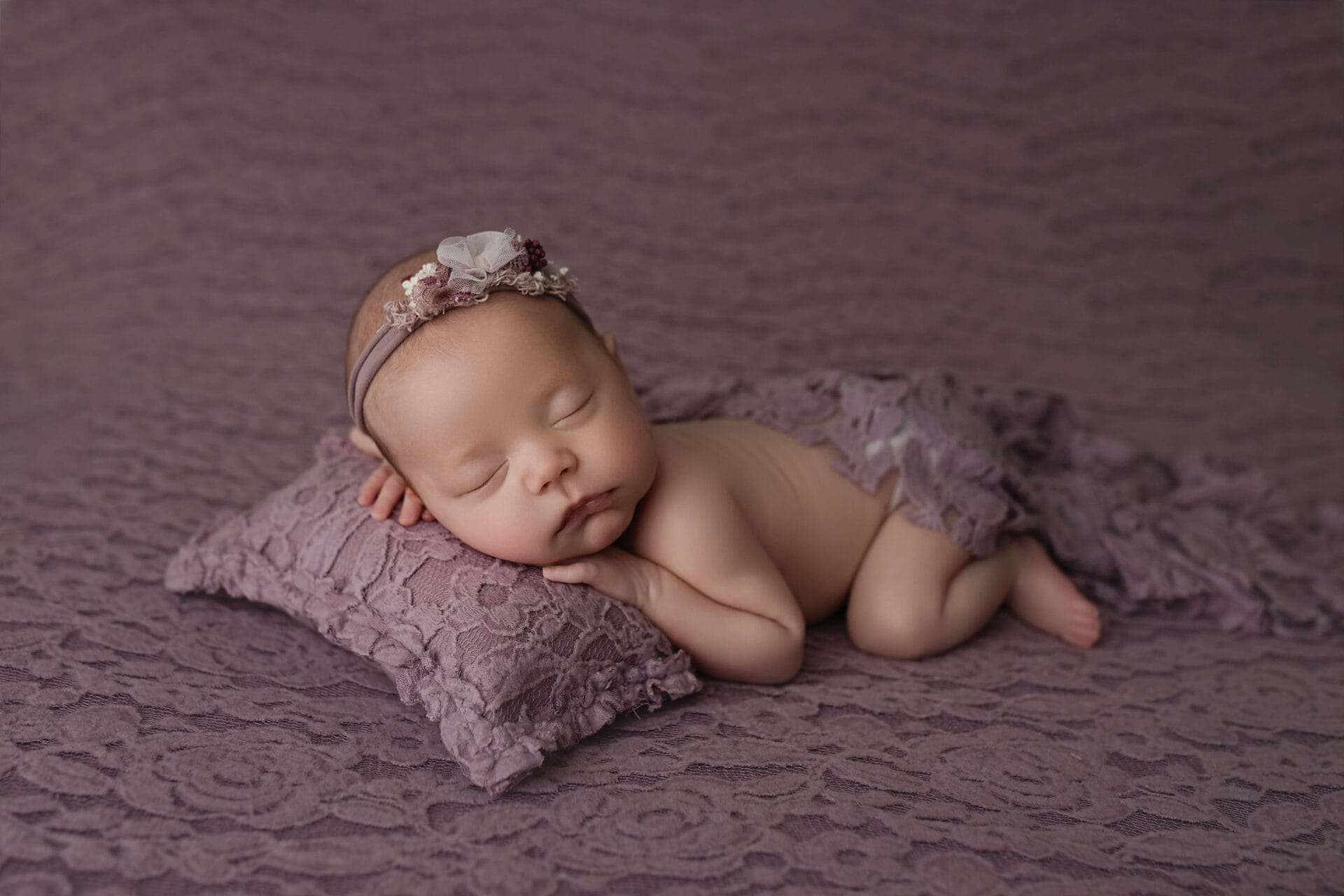 A newborn baby sleeps peacefully on a purple lace pillow, wrapped in a matching purple blanket, and wearing a delicate floral headband. The background and bedding are all a soft lavender color. Melinda Gilmore Photography - Maternity & Newborn Photographer - Louisiana