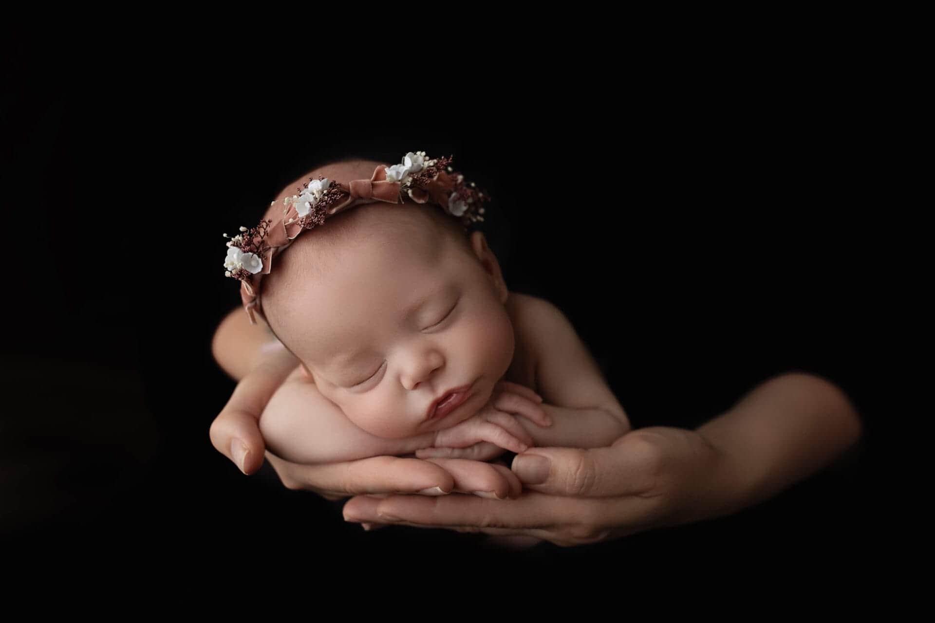 A newborn baby girl posed in her mom's hands with a floral halo in the studio. 