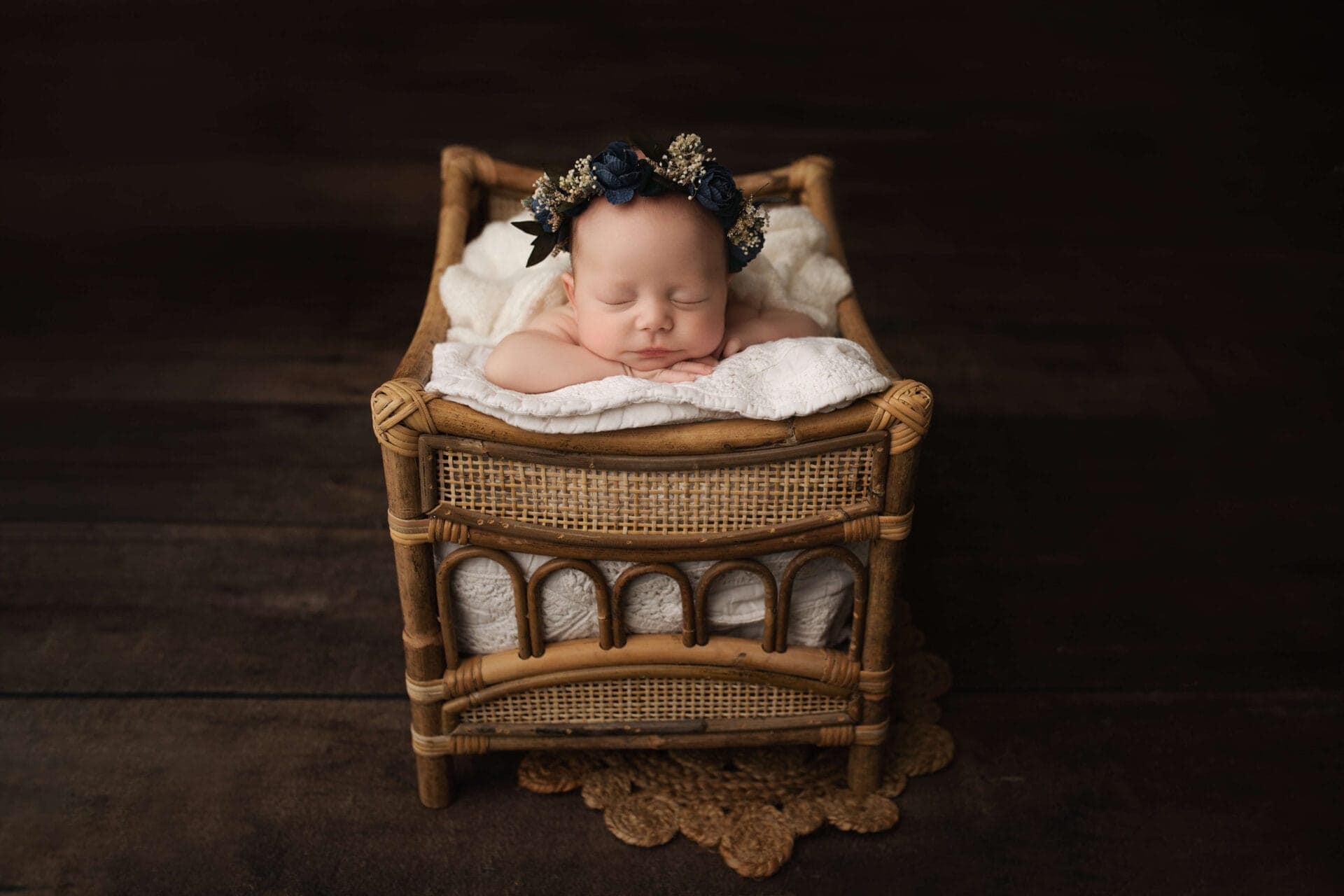 A newborn baby wearing a dark flower headband sleeps peacefully on a white blanket inside a small, vintage-style wicker crib against a dark wooden floor background. Melinda Gilmore Photography - Maternity & Newborn Photographer - Louisiana