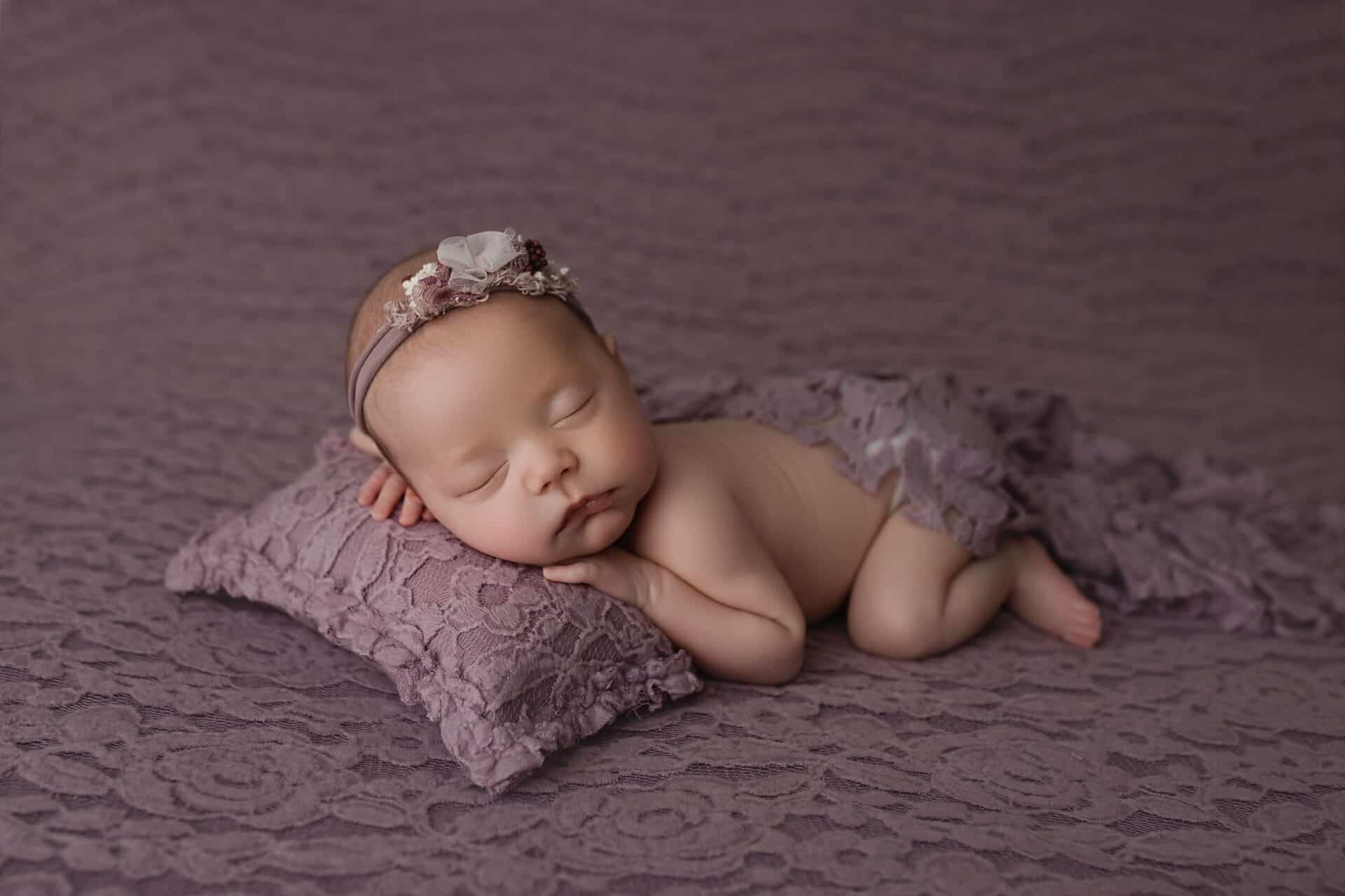 A newborn baby on a lace purple backdrop is lying posed on a pillow wearing a purple headband.