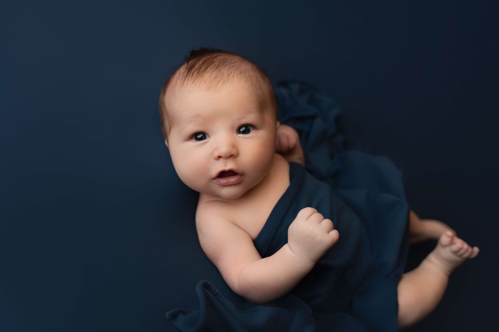 A baby boy for his newborn session in the studio on a blue backdrop looking at the camera.