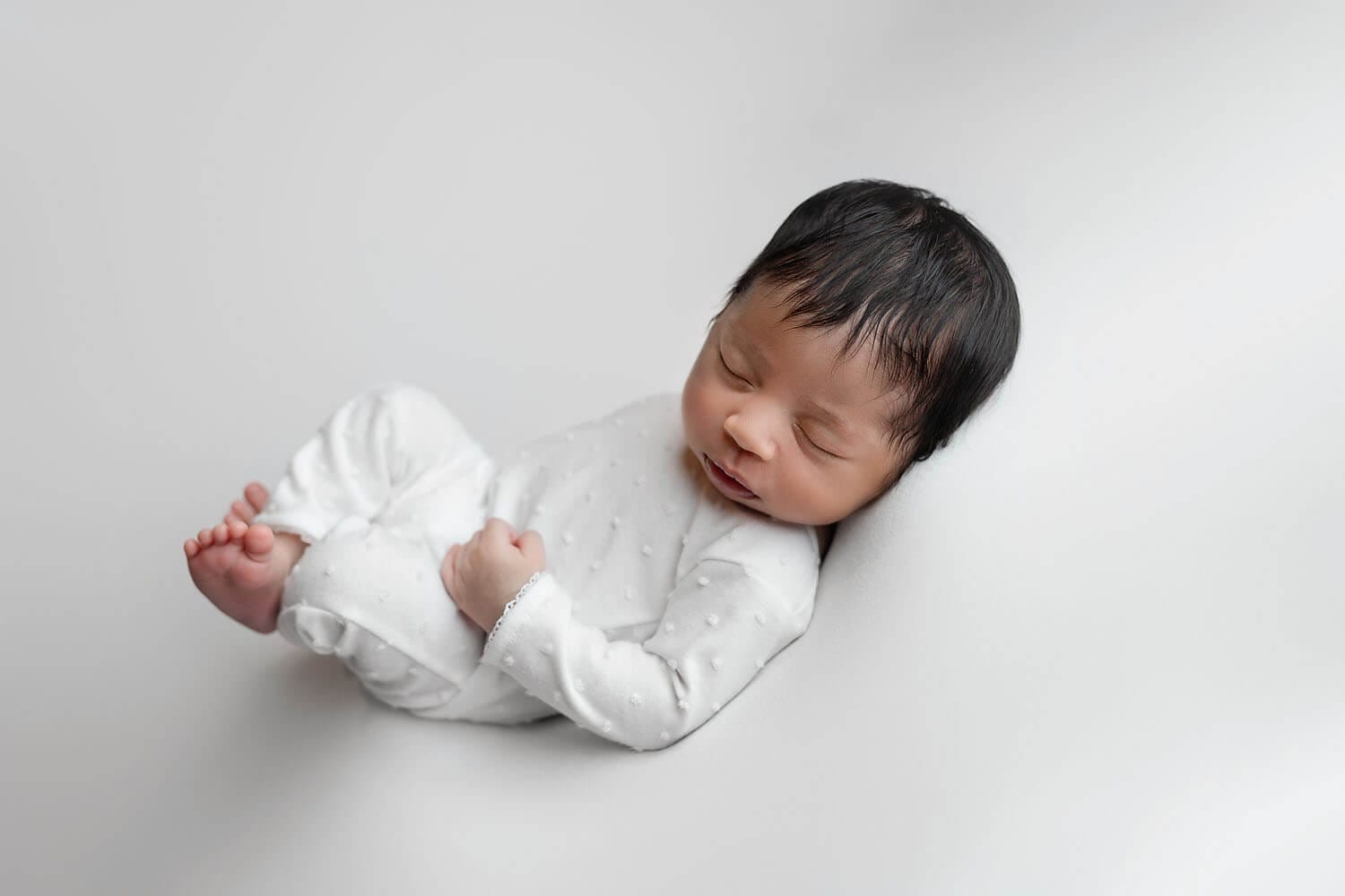A newborn baby lays on a white backdrop wearing a white onesie for newborn photos.