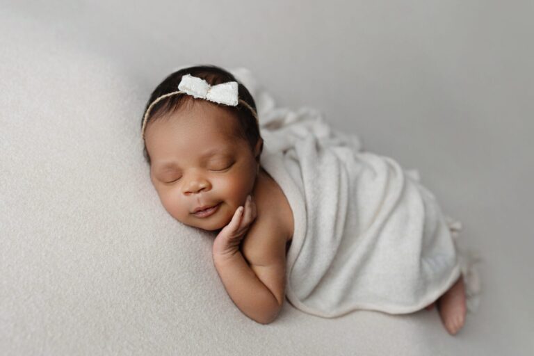 A newborn baby with a white bow headband sleeps peacefully on a soft, light blanket, with one hand resting gently on their cheek. Melinda Gilmore Photography - Maternity & Newborn Photographer - Louisiana
