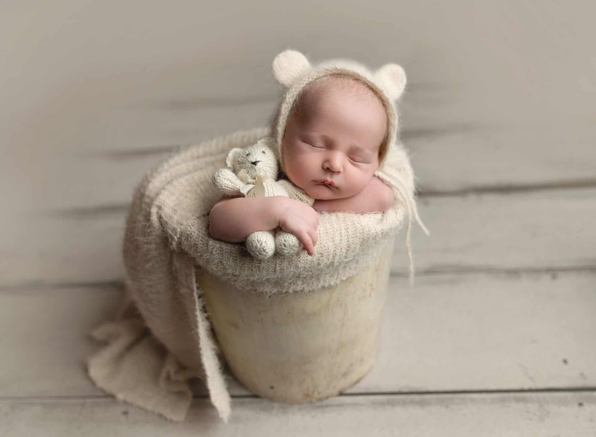 A newborn baby sleeps in a cream-colored bucket, wearing a cozy knit outfit with bear ears and holding a small knit teddy bear, nestled on a soft blanket on a wooden floor. Melinda Gilmore Photography - Maternity & Newborn Photographer - Louisiana