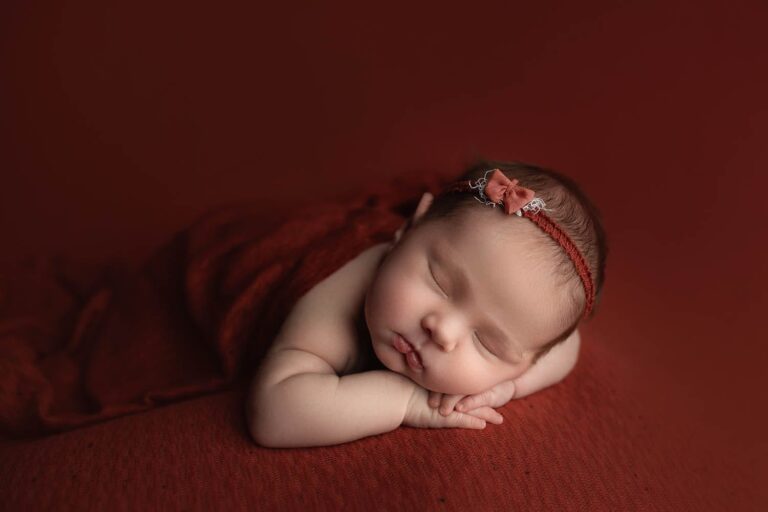 A newborn baby sleeps peacefully on a red blanket, with their head resting on their hands and wearing a red headband with a small bow. The background is also red, creating a warm and serene atmosphere. Melinda Gilmore Photography - Maternity & Newborn Photographer - Louisiana