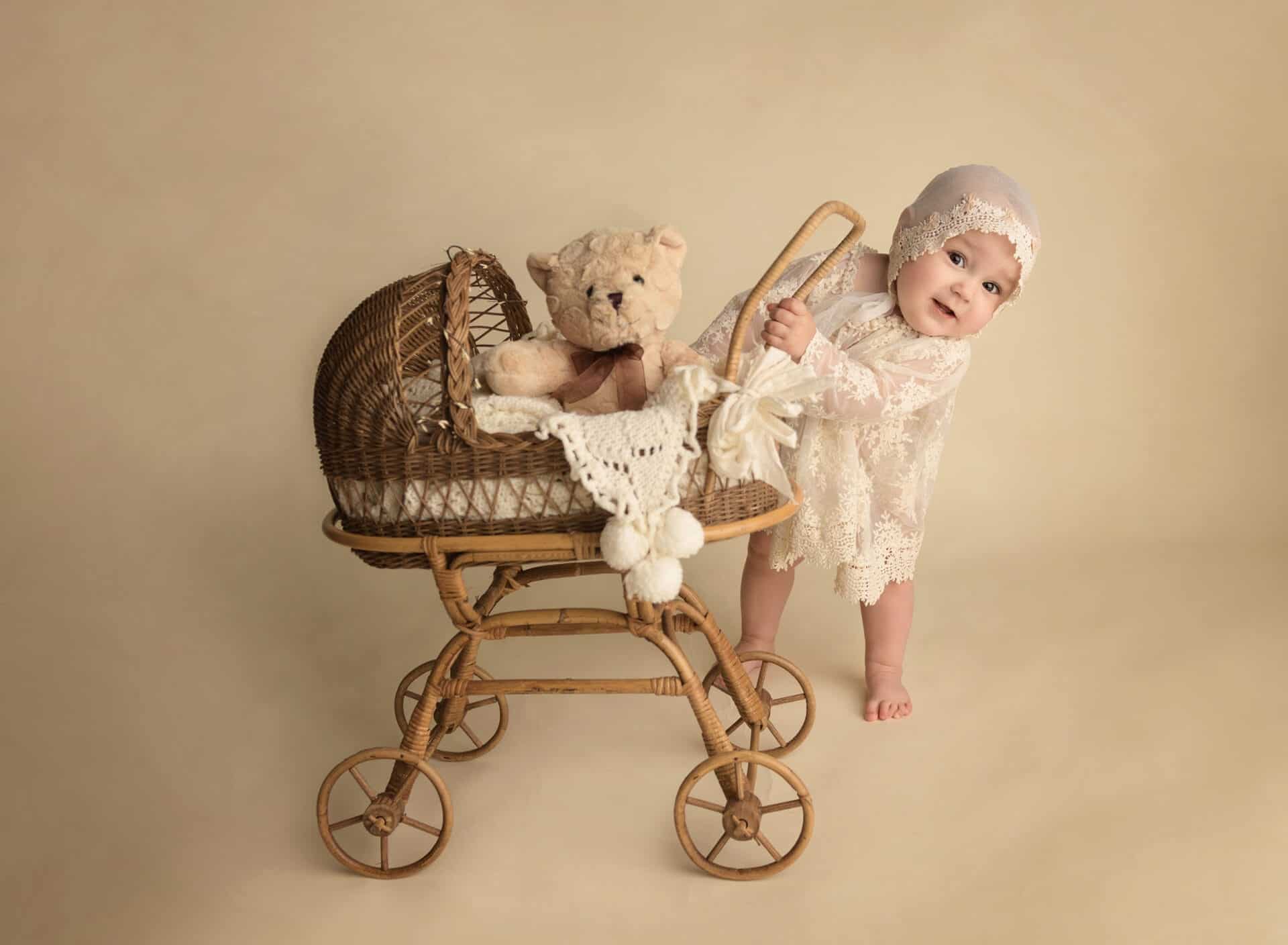 A smiling baby in a lace outfit and bonnet stands beside a vintage wicker stroller, holding its handle. A teddy bear sits inside the stroller, and the background is a soft beige. Melinda Gilmore Photography - Maternity & Newborn Photographer - Louisiana