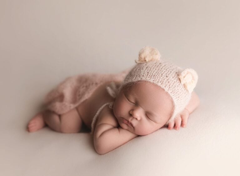A newborn baby sleeps peacefully on a soft surface, wearing a knitted bonnet with small bear ears and a light pink wrap, with one arm tucked under their head. Melinda Gilmore Photography - Maternity & Newborn Photographer - Louisiana