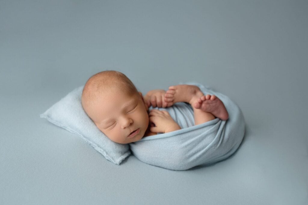 A newborn baby is peacefully sleeping on a small pillow, wrapped snugly in a light blue blanket, against a matching soft blue background. Melinda Gilmore Photography - Maternity & Newborn Photographer - Louisiana