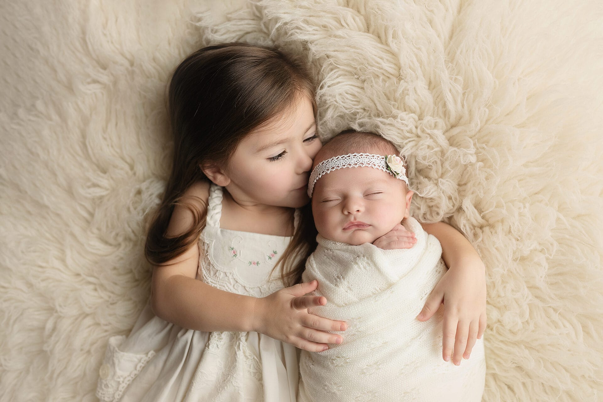 A new big sister from New Orleans is in the studio with her baby sister for her newborn session, lying in her arms and kissing her.