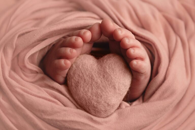 A close-up of a baby’s tiny feet gently holding a soft, pink, heart-shaped object, surrounded by a pink fabric wrap. Melinda Gilmore Photography - Maternity & Newborn Photographer - Louisiana