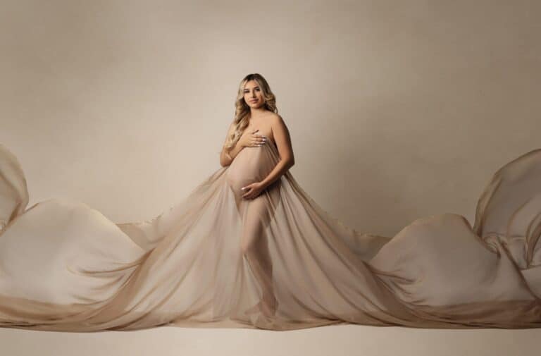A pregnant woman stands barefoot against a beige background, wearing a flowing, sheer nude fabric that drapes around her body and extends outward. She cradles her belly with one hand and rests the other on her chest, looking serene. Melinda Gilmore Photography - Maternity & Newborn Photographer - Louisiana