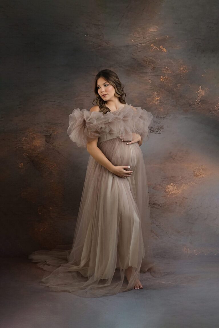 A pregnant woman in a flowing, sheer beige gown stands barefoot against a softly lit, artistic studio backdrop, gently cradling her belly and looking down with a serene expression. Melinda Gilmore Photography - Maternity & Newborn Photographer - Louisiana
