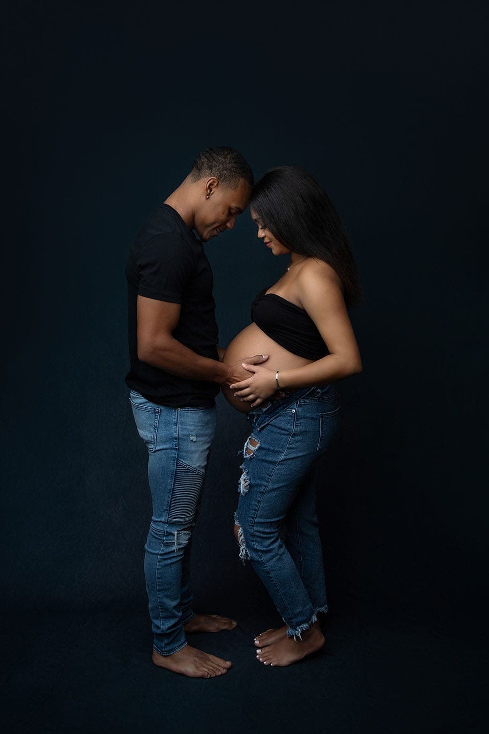 A couple stands barefoot facing each other against a dark background, both wearing black tops and jeans. The man gently touches the woman's pregnant belly, and they both smile warmly. Melinda Gilmore Photography - Maternity & Newborn Photographer - Louisiana