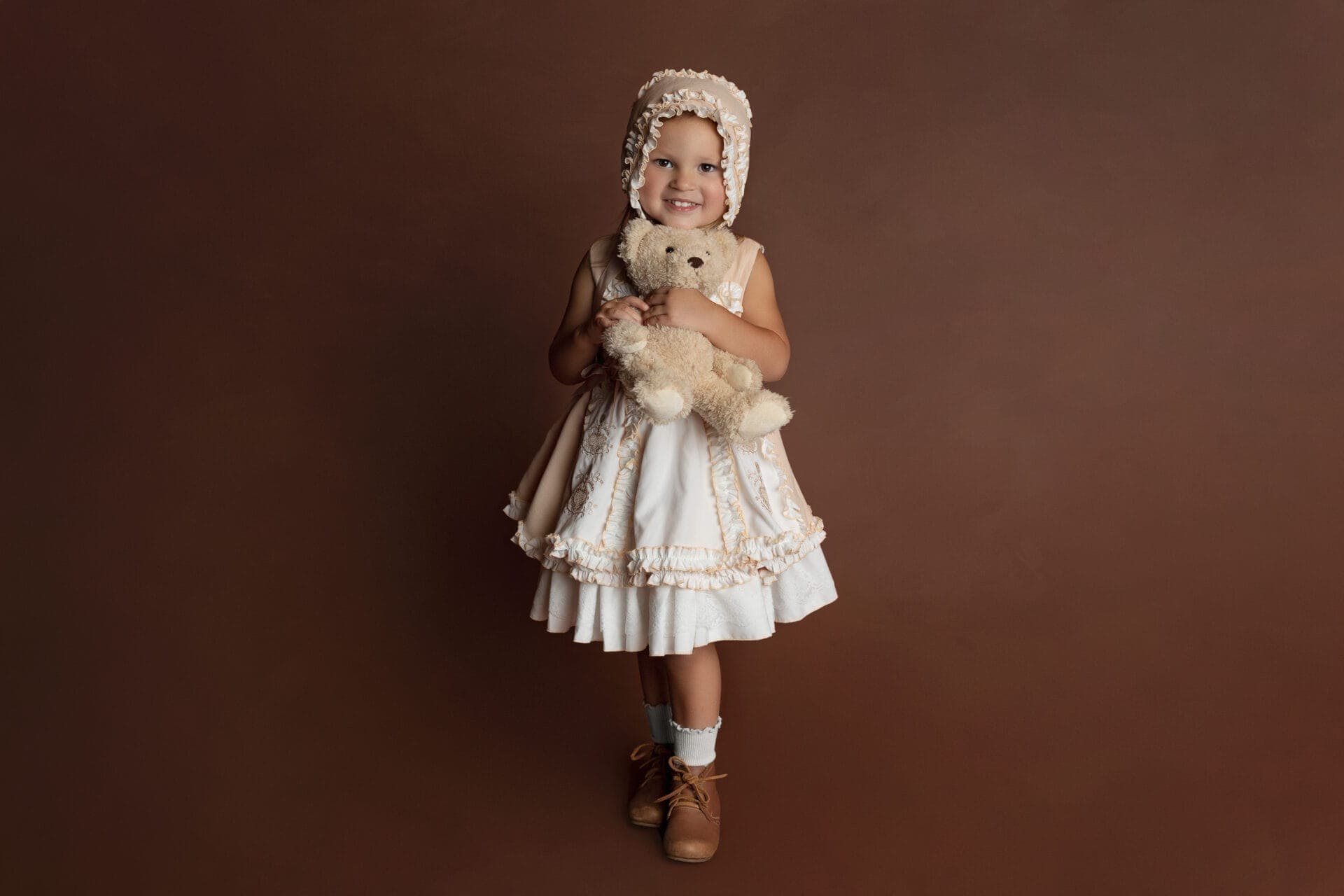 A young child in a vintage cream dress and bonnet smiles while hugging a teddy bear, standing against a plain brown background. Melinda Gilmore Photography - Maternity & Newborn Photographer - Louisiana
