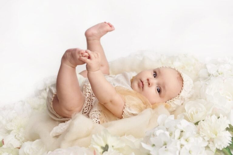 A baby in a cream outfit and bonnet lies on a soft, fluffy blanket surrounded by white flowers, holding its feet and looking up with a calm expression. Melinda Gilmore Photography - Maternity & Newborn Photographer - Louisiana