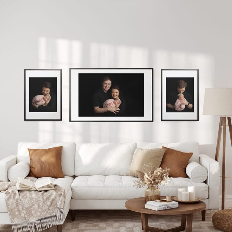 A white sofa with brown and tan cushions sits beneath three framed family photos on a wall; a man and woman hold a newborn in the center photo, with individual portraits on either side. A lamp and coffee table complete the cozy room. Melinda Gilmore Photography - Maternity & Newborn Photographer - Louisiana