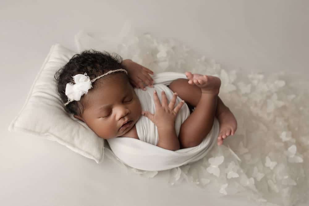 A sleeping baby wrapped in a white blanket lies on a white pillow, with a white flower headband in her hair. Soft white petals or tulle surround her, creating a peaceful and gentle scene. Melinda Gilmore Photography - Maternity & Newborn Photographer - Louisiana
