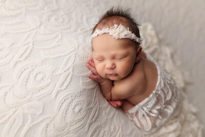 A newborn baby sleeps peacefully on a soft white textured blanket, wrapped in a white lace cloth with a matching lace headband. The baby’s arms are tucked close, and the scene looks calm and serene. Melinda Gilmore Photography - Maternity & Newborn Photographer - Louisiana
